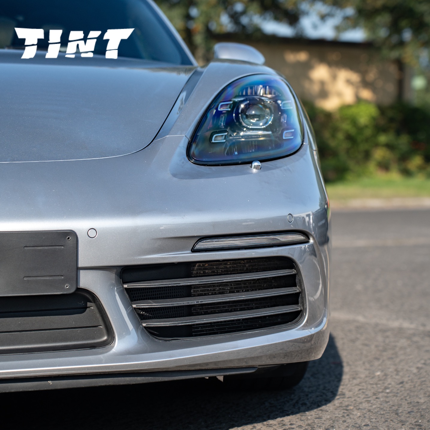 Szary Porsche, zbliżenie na przedni reflektor z widocznym napisem 'TINT' na szybie. Detal maski, grilla i zderzaka.