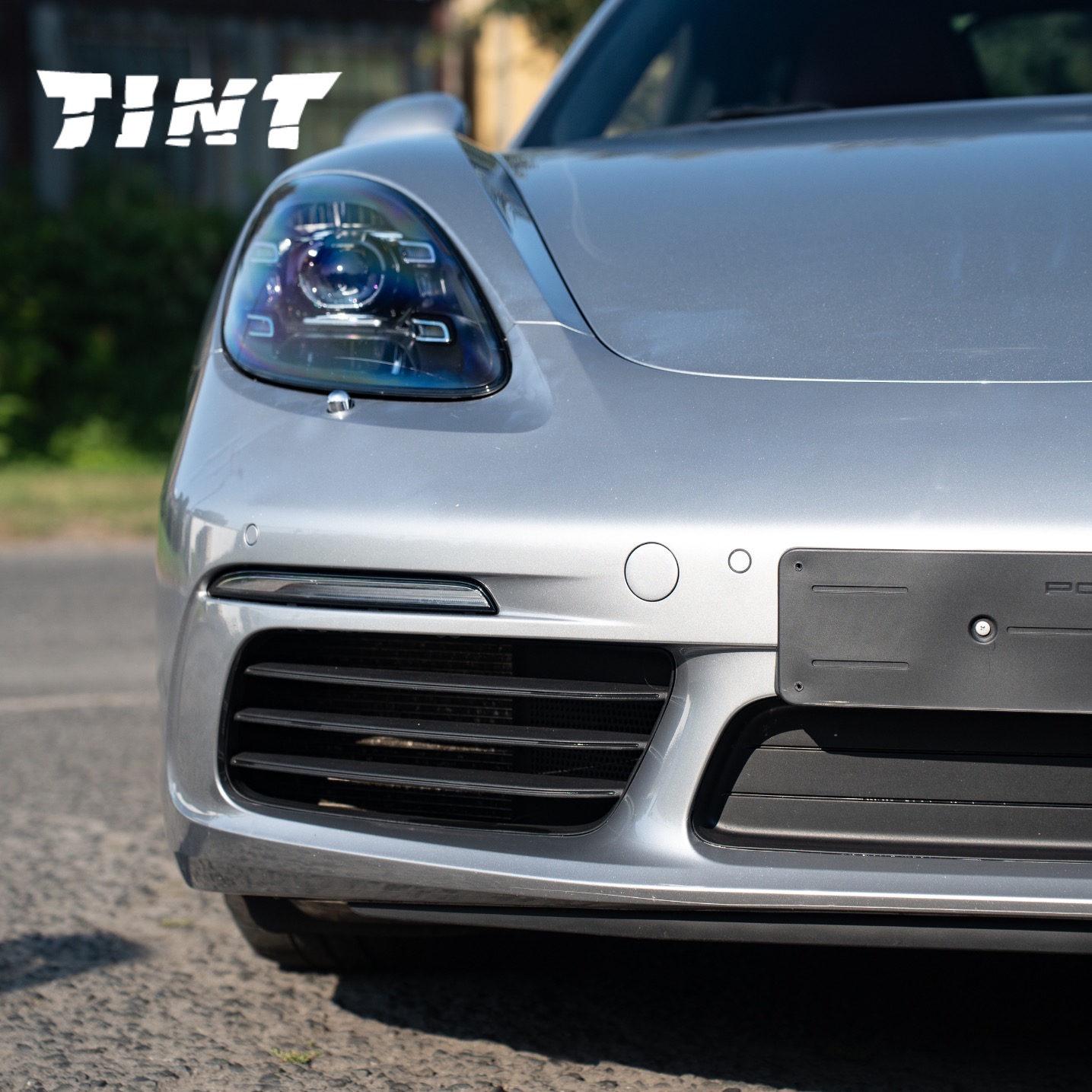 Szary Porsche Panamera z bliska, z widocznym logo 'TINT' w lewym górnym rogu, sugerującym przyciemnianie szyb lub oklejanie folią.