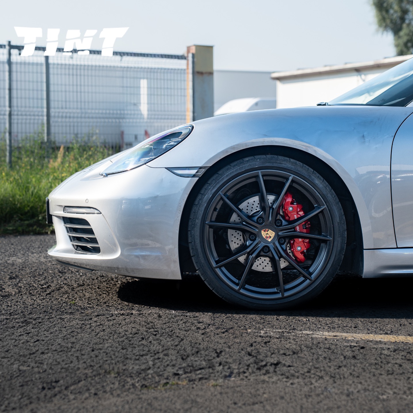 Srebrny Porsche z czarnymi felgami i czerwonymi zaciskami hamulcowymi na tle parkingu i ogrodzenia. Widoczny fragment logo firmy.