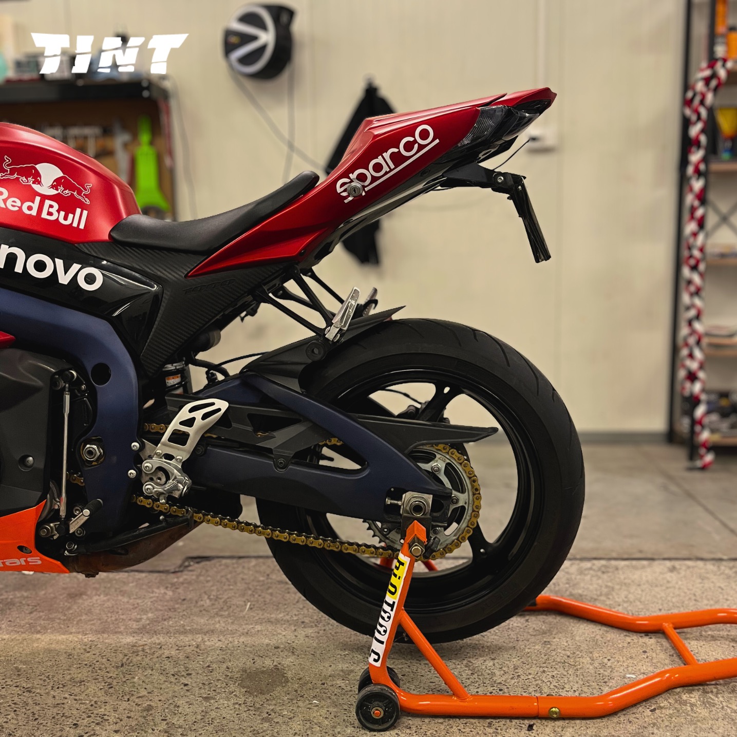 Sportowy motocykl z naklejkami Red Bull i Sparco, ustawiony na pomarańczowym stojaku warsztatowym, widok z boku na tylne koło i detale graficzne.