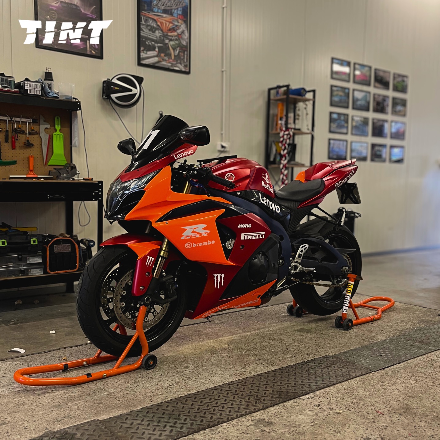 Pomarańczowo-czerwony motocykl sportowy z naklejkami sponsorskimi, umieszczony na pomarańczowych stojakach w garażu. Widoczne narzędzia i plakaty na ścianie.