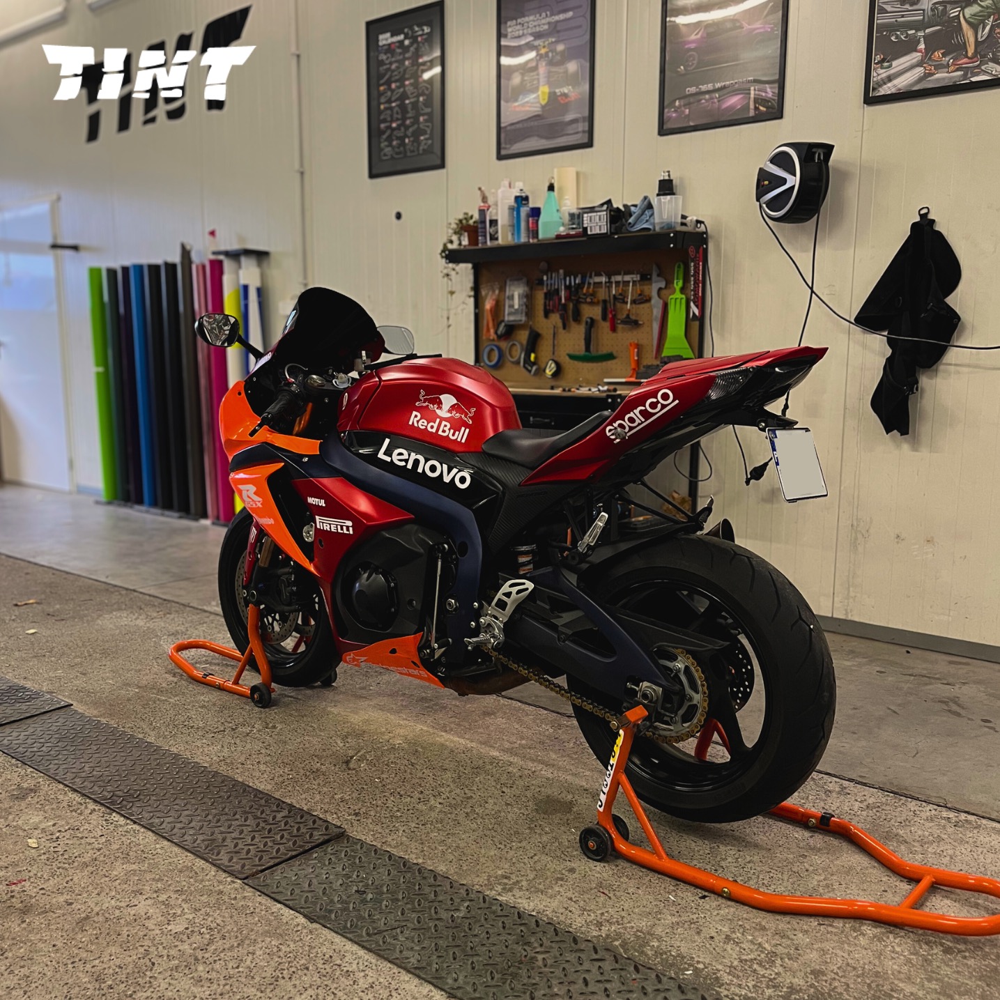 Oklejony motocykl sportowy w barwach Red Bulla i Lenovo, stojący w warsztacie z rolkami folii i narzędziami. Widoczne logo firmy 'TINT' na ścianie.