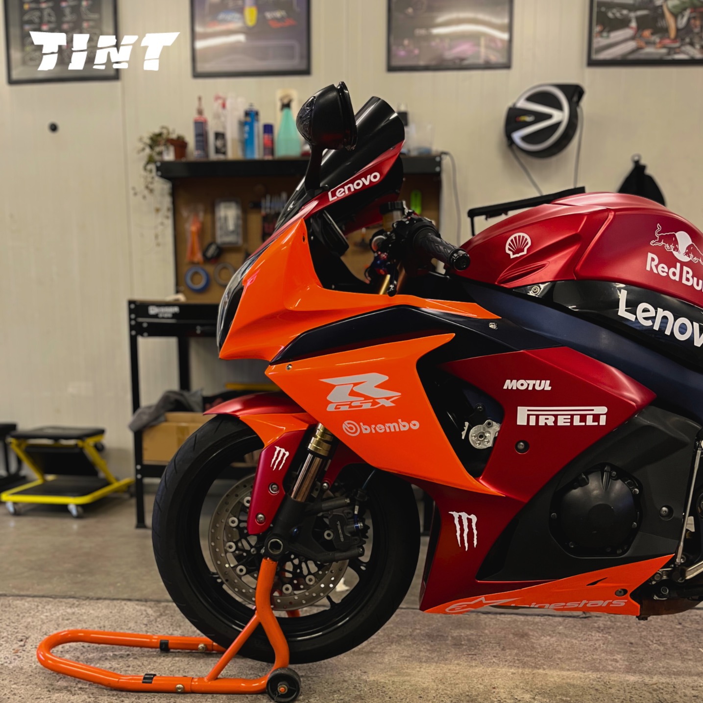 Oklejony motocykl Suzuki GSX-R w barwach Red Bull Lenovo z logo Pirelli i Motul, na pomarańczowym stojaku, w warsztacie.