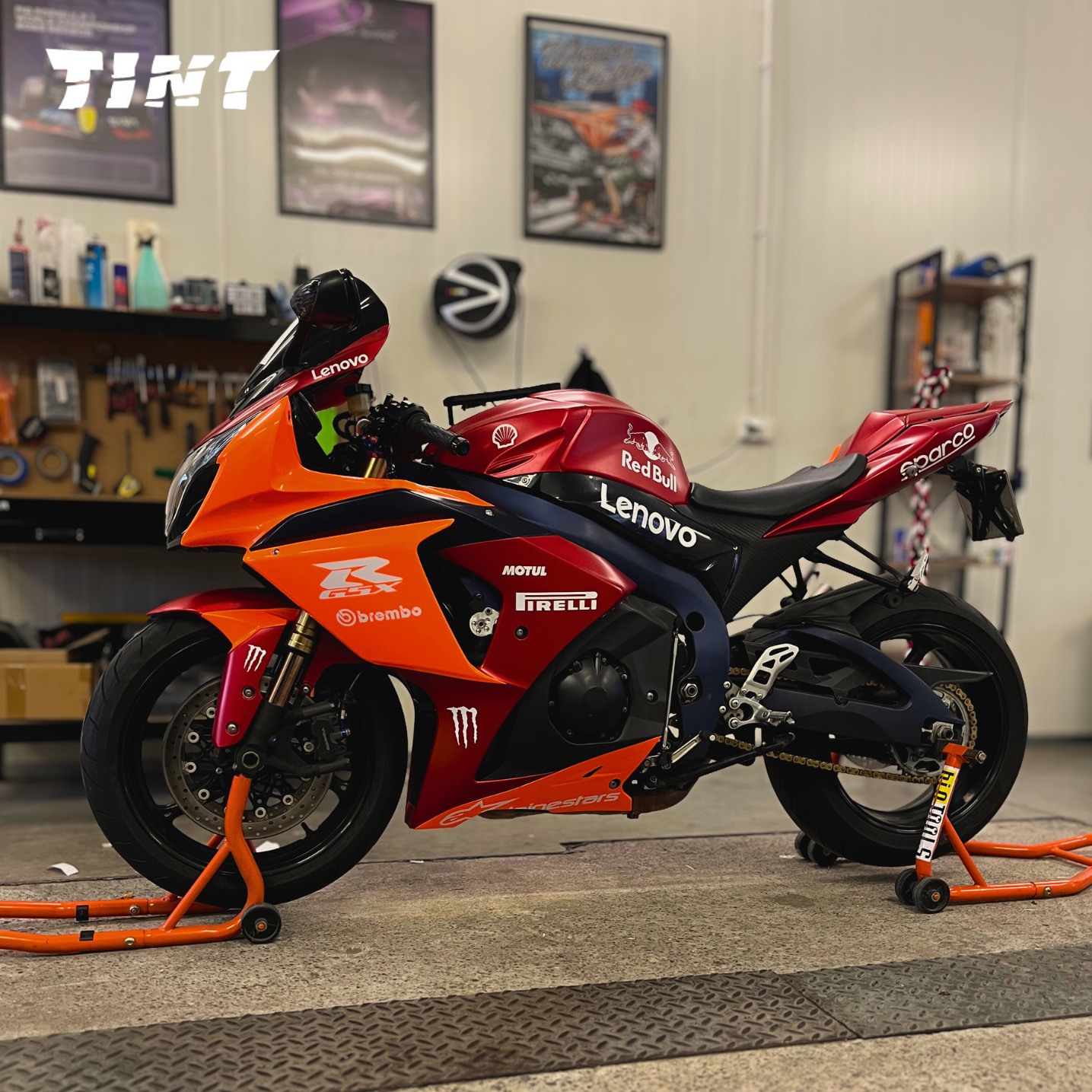 Oklejony motocykl sportowy w barwach Red Bull i Lenovo, z widocznymi logotypami Pirelli, Brembo i Alpinestars, stojący w warsztacie na pomarańczowych stojakach.