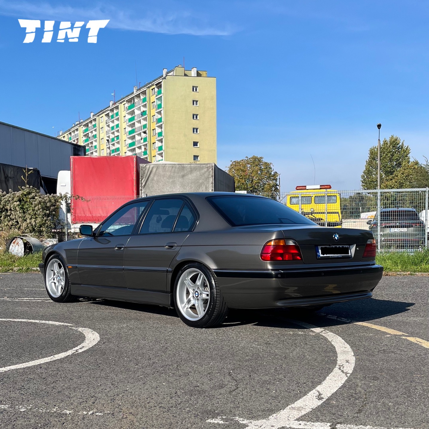 Szary samochód BMW serii 7 (E38) z aluminiowymi felgami na parkingu, z budynkiem mieszkalnym w tle. Widoczny napis 'TINT' w lewym górnym rogu.