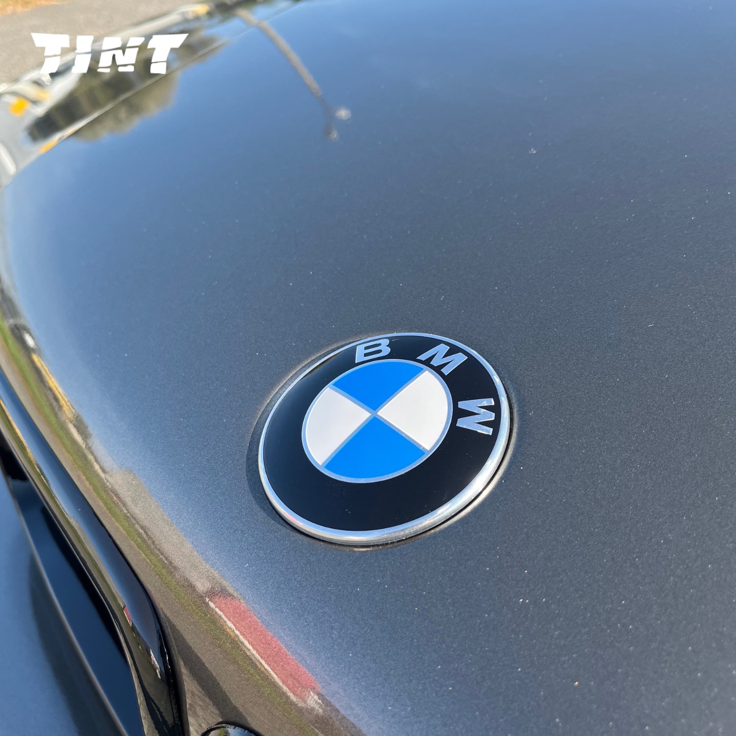Zbliżenie na maskę szarego BMW z wyraźnym logo marki. Oświetlenie słoneczne podkreśla detale lakieru. Widoczny fragment karoserii.