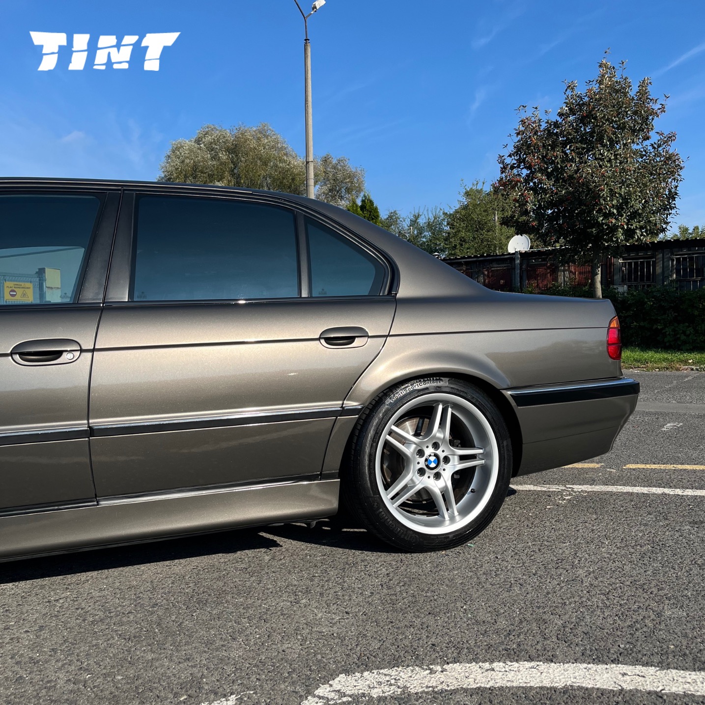 Przyciemnione szyby w brązowym BMW serii 7 E38, zaparkowanym na asfalcie. Widoczne logo firmy TINT w lewym górnym rogu. Srebrne felgi.