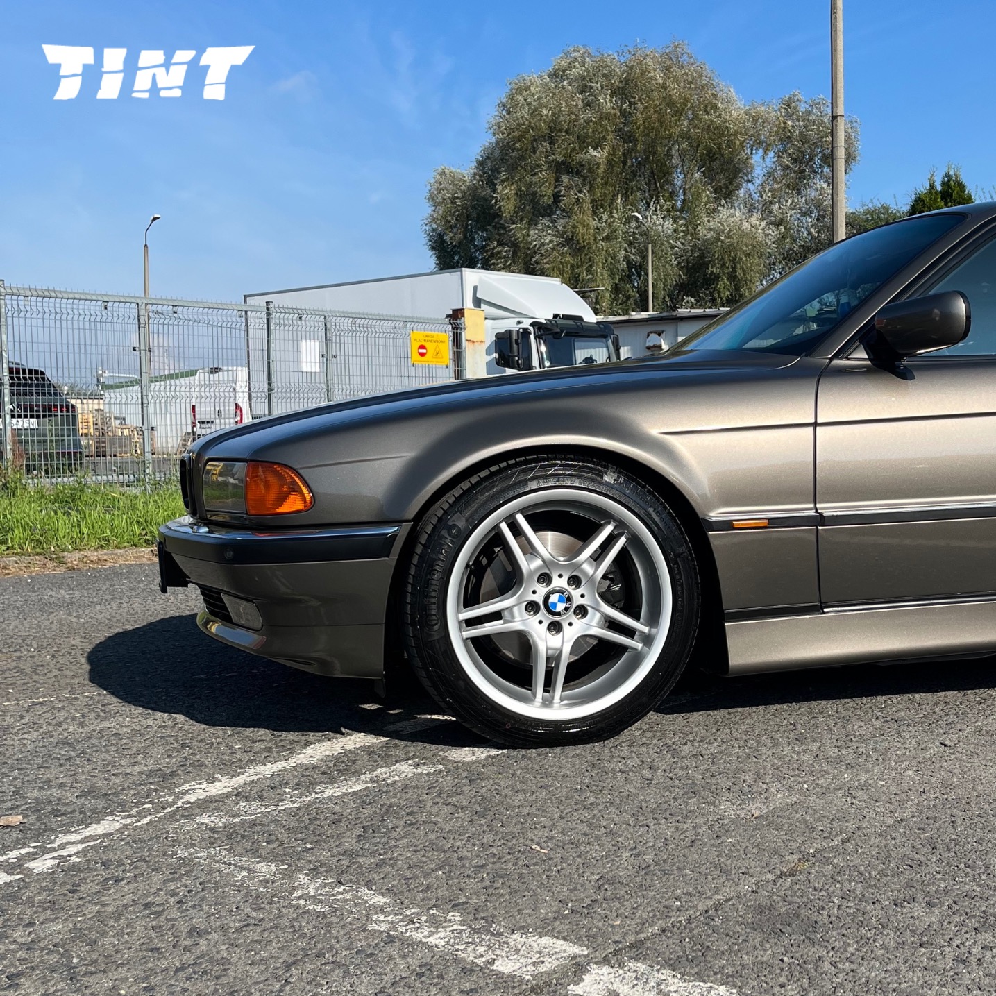 Szaro-złoty samochód BMW serii 7 z aluminiowymi felgami na parkingu, widok z boku, z logo firmy TINT w lewym górnym rogu. Słoneczny dzień.