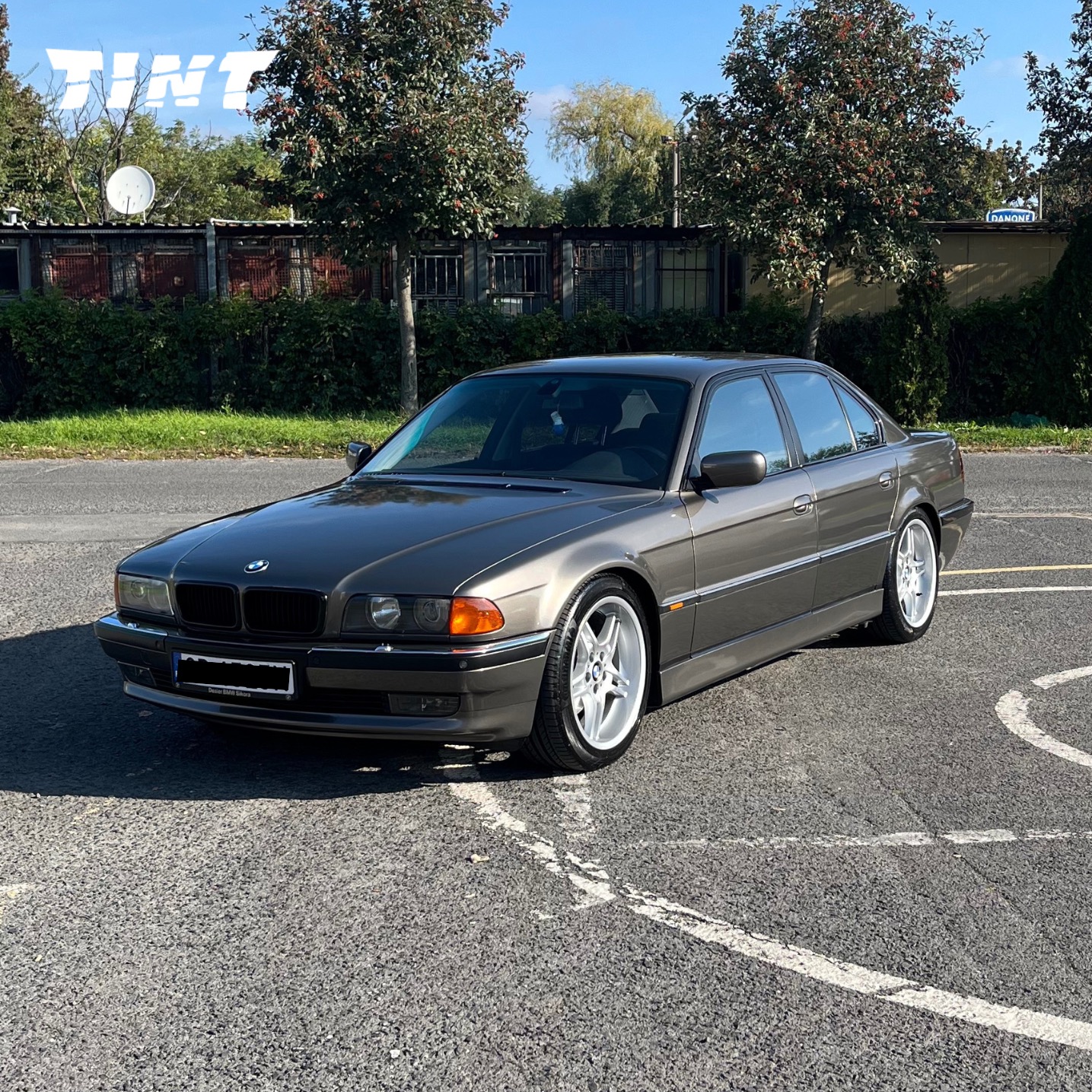 Szary BMW serii 7 (E38) z aluminiowymi felgami na parkingu w słoneczny dzień. Widoczne logo firmy na szybie, sugerujące przyciemnianie szyb.