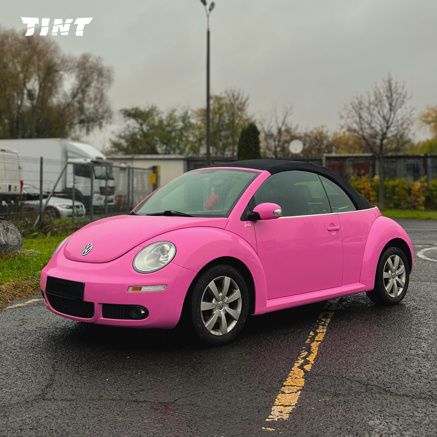 Różowy Volkswagen Beetle Cabrio po zmianie koloru, zaparkowany na mokrym asfalcie. Widoczne logo firmy w lewym górnym rogu. Auto w stylu Barbie.