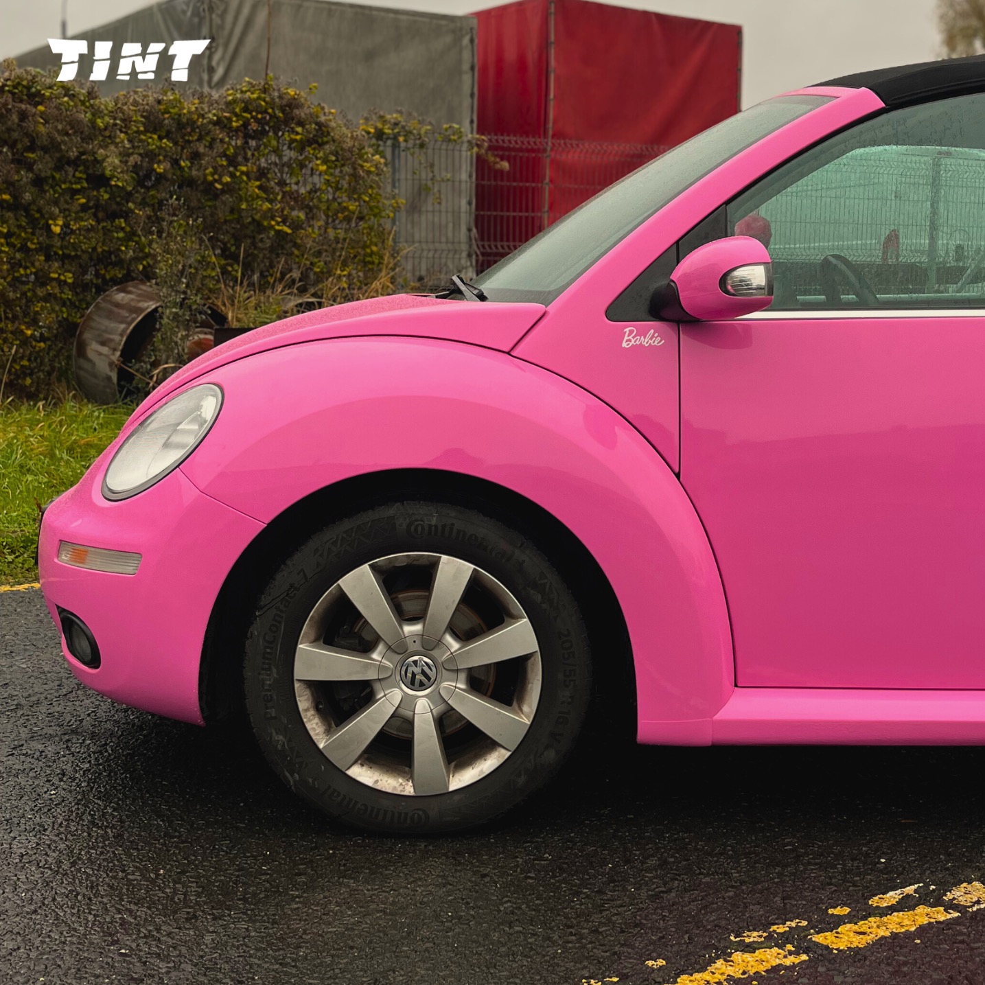 Różowy Volkswagen Beetle Cabrio z napisem 'Barbie' na błotniku, zaparkowany na mokrym asfalcie z żółtą linią. Widoczne logo firmy na szybie.