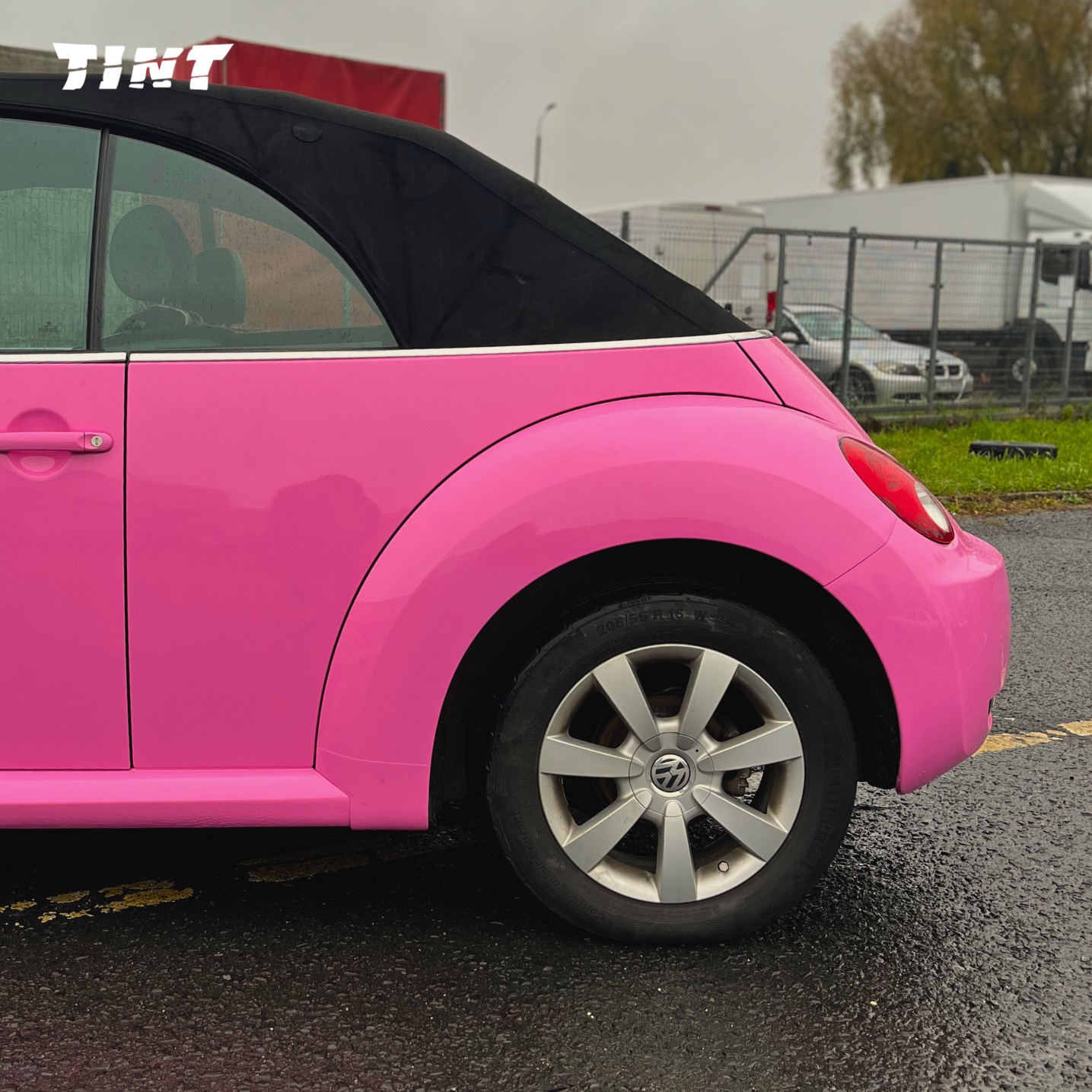 Różowy kabriolet Volkswagen Beetle z czarnym dachem, widziany z boku na mokrym asfalcie. W tle ogrodzenie i białe samochody dostawcze.