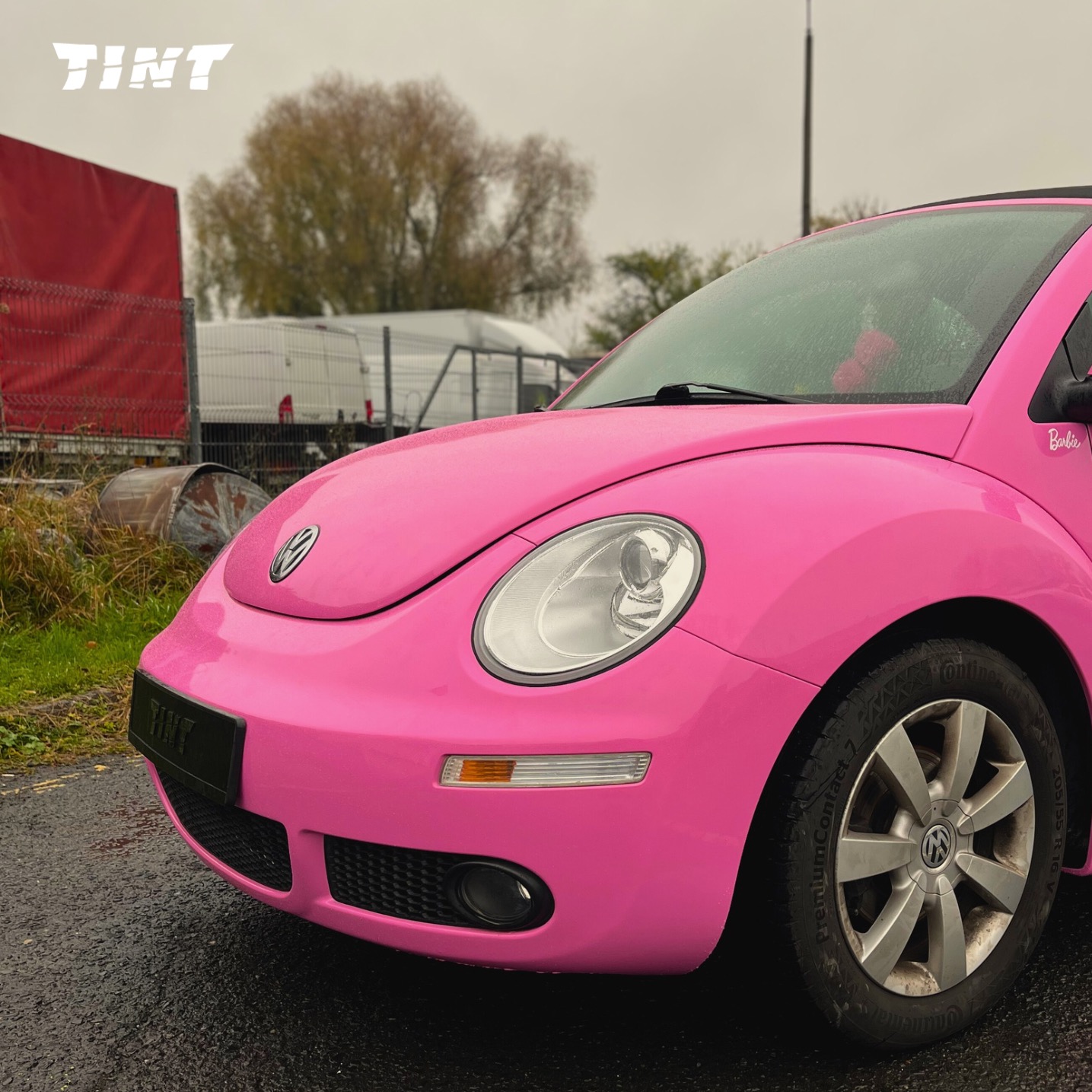 Różowy Volkswagen Beetle z naklejką 'Barbie' na błotniku, zaparkowany na mokrym asfalcie. Widoczne logo firmy na tablicy rejestracyjnej.