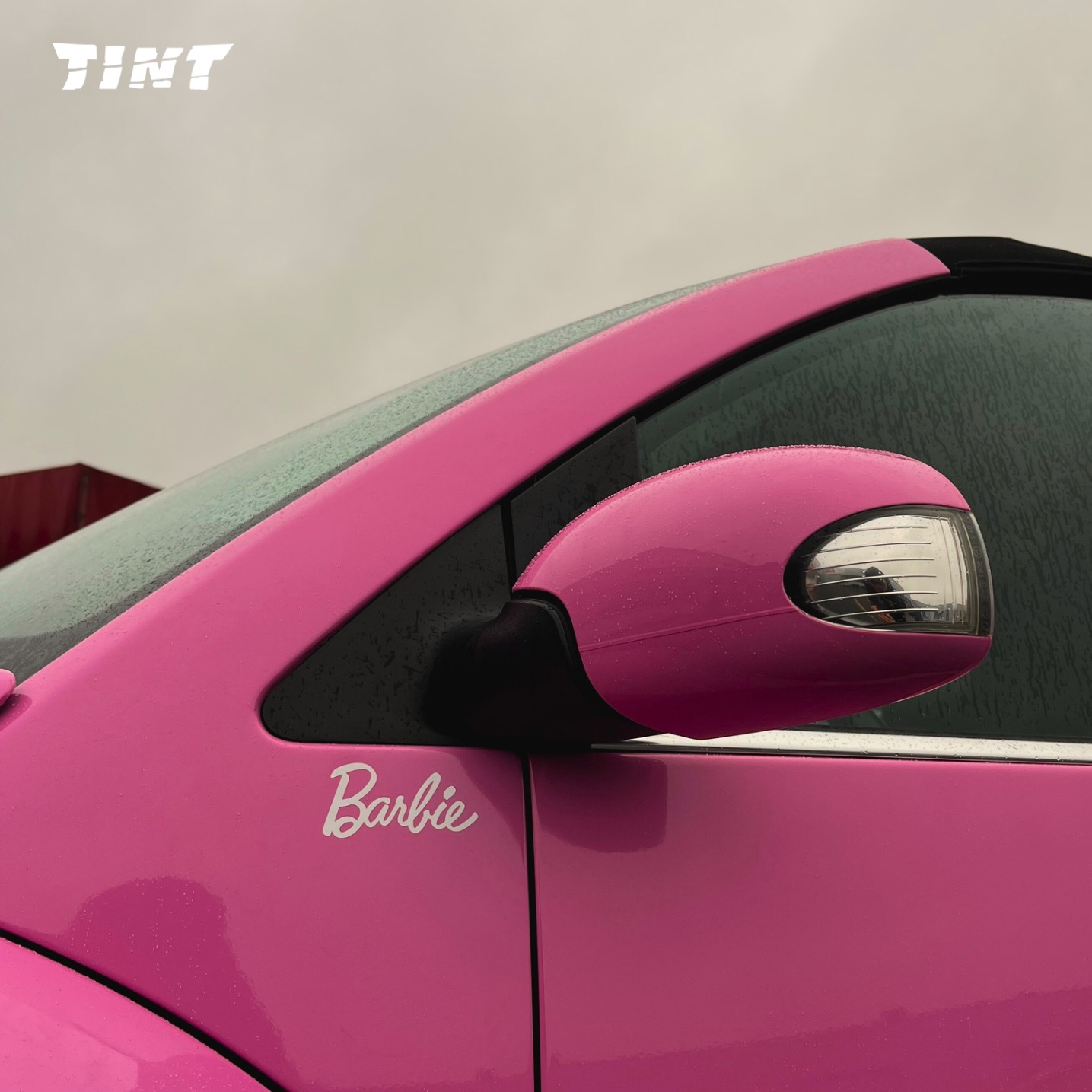 Różowy samochód z napisem 'Barbie' na drzwiach, po oklejeniu folią. Widoczne lusterko i fragment szyby z efektem przyciemnienia. Deszczowa pogoda.