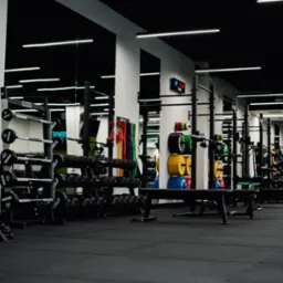 Profesjonalna siłownia z rzędami hantli, sztang, obciążników i gum oporowych, z zegarem elektronicznym na ścianie.