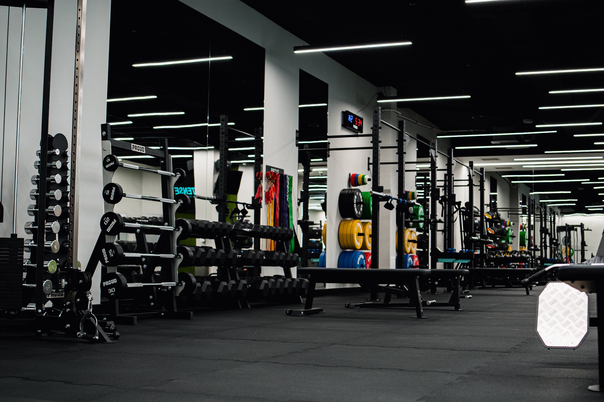 Profesjonalna siłownia z rzędami hantli, sztang, obciążników i gum oporowych, z zegarem elektronicznym na ścianie.