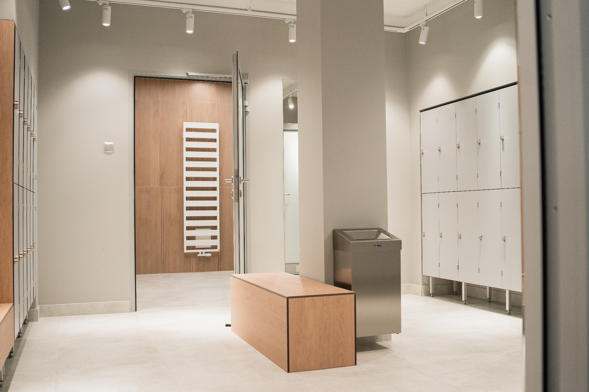 Jasne wnętrze szatni z drewnianymi i białymi szafkami, metalowym koszem na śmieci i drewnianą ławką, oświetlone punktowymi lampami na suficie.