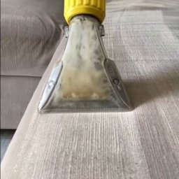 CleanGlow&Dynasty - Dysza odkurzacza piorącego z żółtą obudową czyszcząca jasnoszarą tapicerkę sofy, widoczne ślady wilgoci i zabrudzeń wewnątrz przezroczystej końcówki.