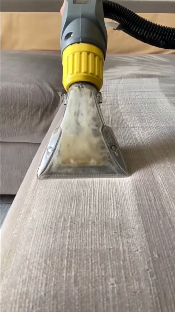 Dysza odkurzacza piorącego z żółtą obudową czyszcząca jasnoszarą tapicerkę sofy, widoczne ślady wilgoci i zabrudzeń wewnątrz przezroczystej końcówki.