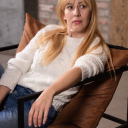 NO.TAK.STUDIO Natalia Olszewska - Portret kobiety z blond włosami w białym swetrze, siedzącej w brązowym fotelu na tle ceglanej ściany.
