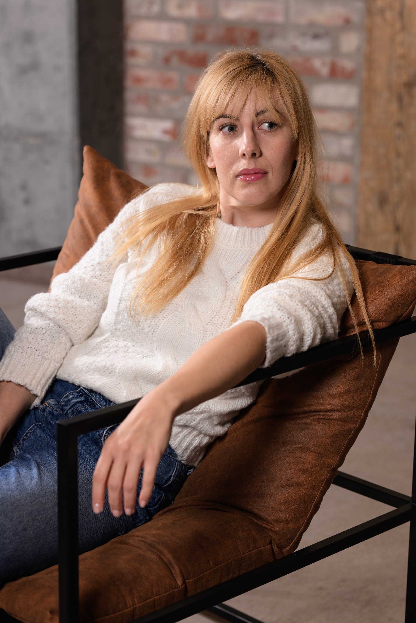 Portret kobiety z blond włosami w białym swetrze, siedzącej w brązowym fotelu na tle ceglanej ściany.