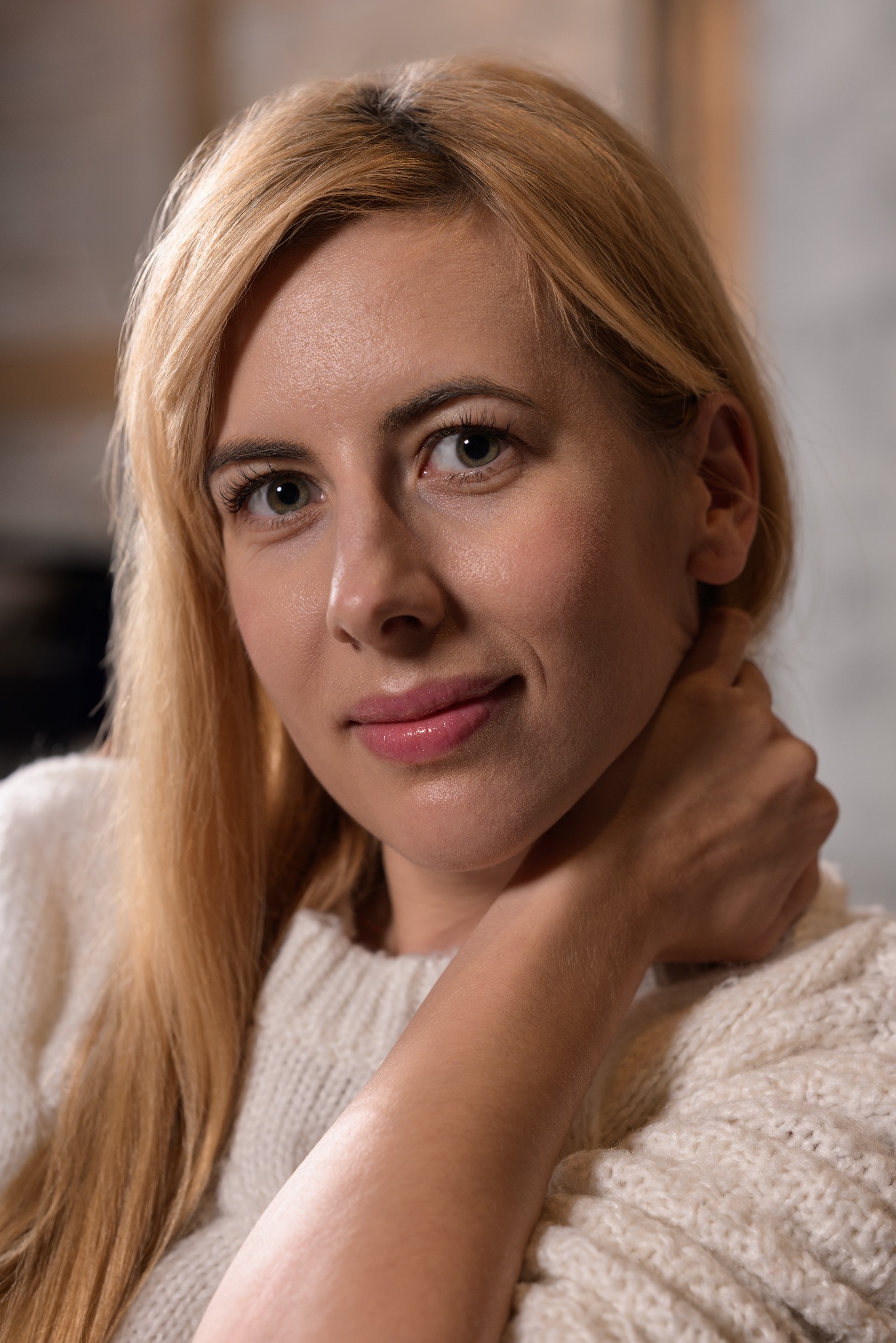 Portret uśmiechniętej blondynki w białym swetrze, z dłonią wspartą na szyi, w neutralnym oświetleniu.