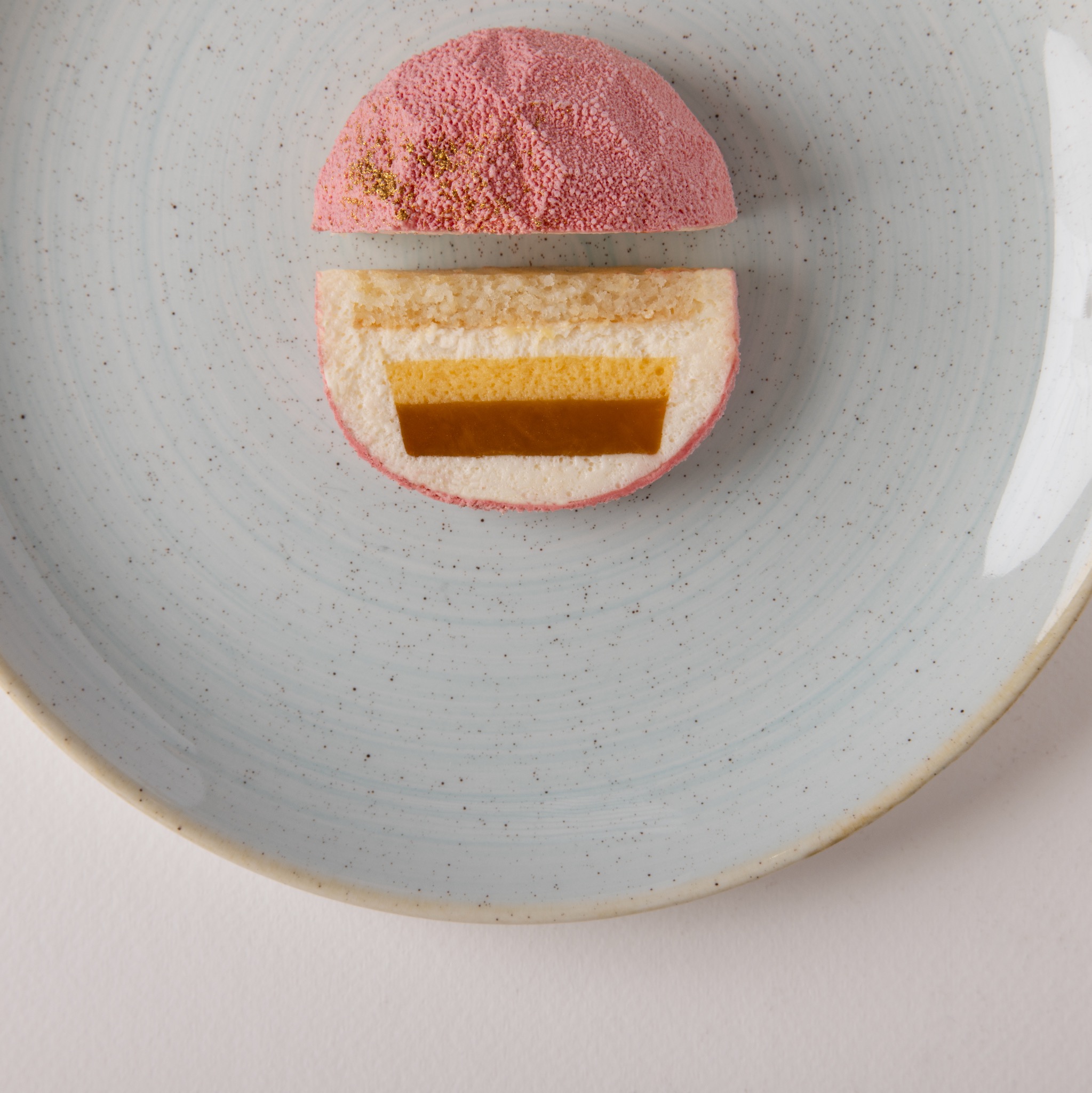 Przekrój różowego deseru z trzema warstwami na jasnoniebieskim talerzu z widocznymi drobnymi kropkami złota na górnej części.
