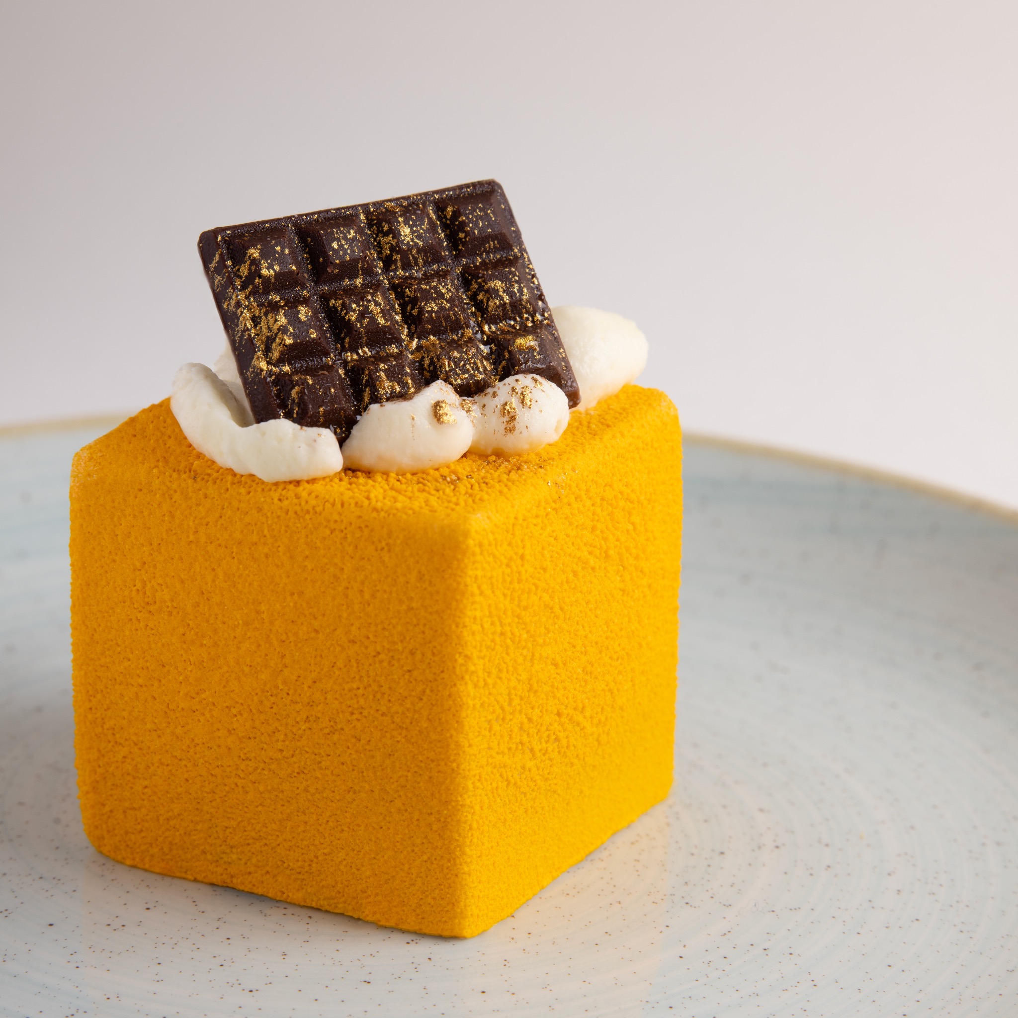 Żółty deser w kształcie kostki z czekoladową dekoracją posypaną złotem i kremowymi kleksami na talerzu.