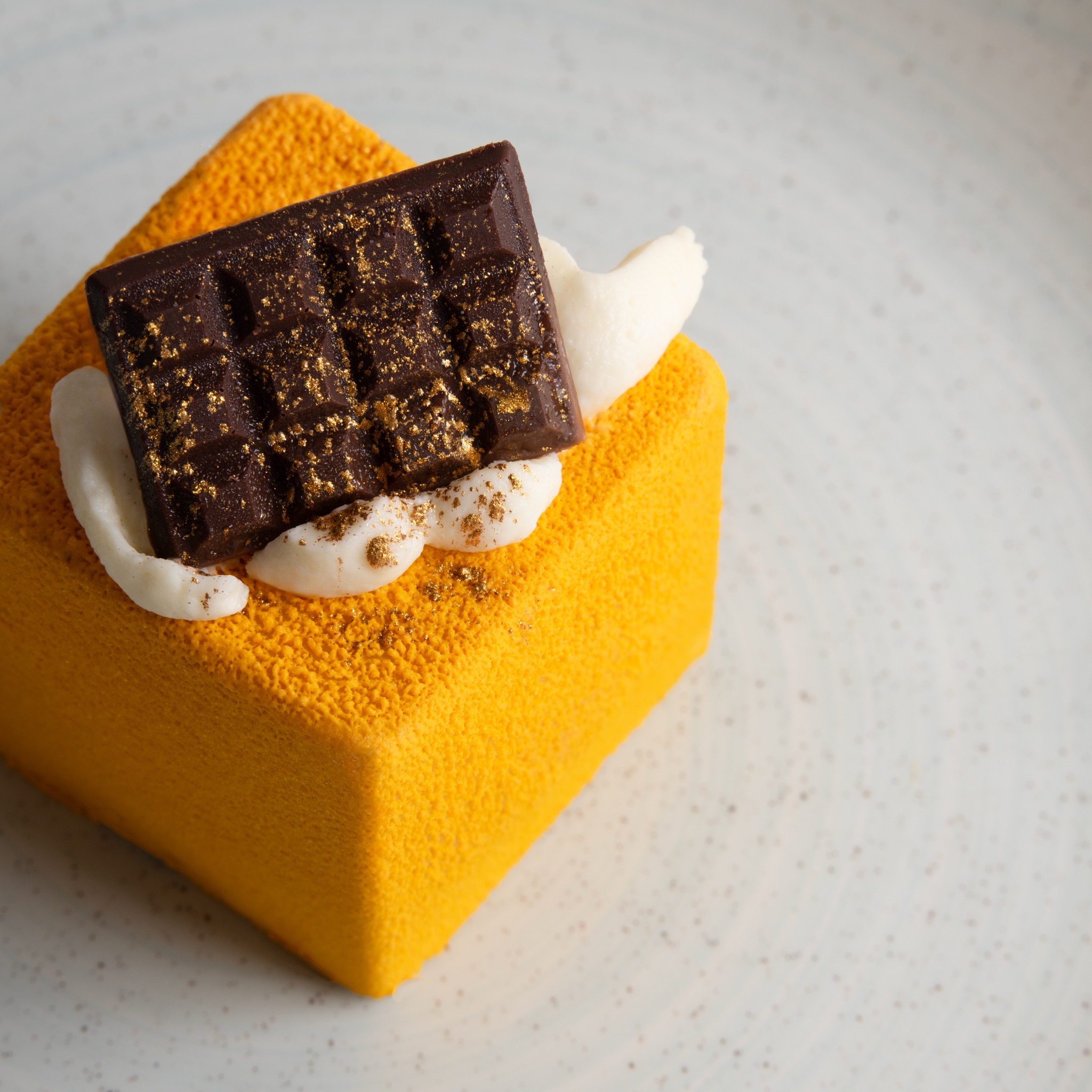 Kwadratowy deser o żółtej, teksturowanej powierzchni, udekorowany czekoladową tabliczką posypaną złotem i kleksami kremu, na jasnym talerzu.