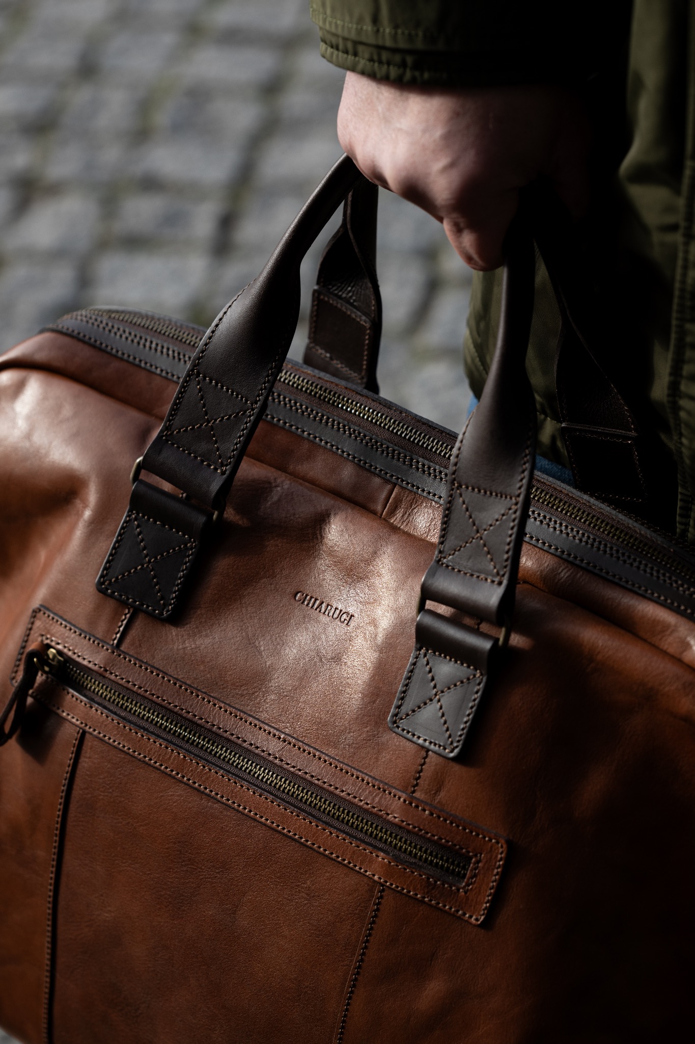Brązowa skórzana torba podróżna marki Chiarugi, trzymana w dłoni na tle chodnika.