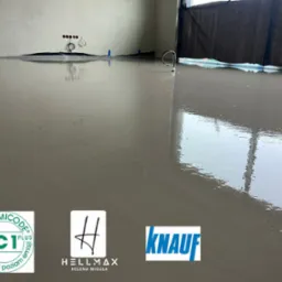 Świeżo wylana, gładka i połyskująca posadzka w jasnym odcieniu szarości, z widocznymi przewodami wychodzącymi ze ściany i logotypami producentów materiałów budowlanych: GEV-EMICODE, Hellmax i Knauf.