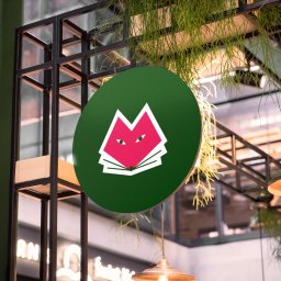 Maryna Slobodianiuk - Zielony okrągły szyld z różowym logo w kształcie liska nad otwartą książką, wiszący pod sufitem z dekoracyjnymi roślinami i oświetleniem.