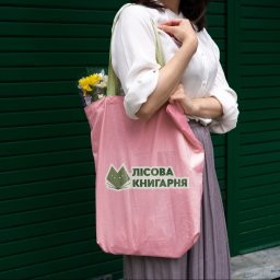 Maryna Slobodianiuk - Kobieta trzyma różową torbę z nadrukowanym logo w kształcie kota i napisem w języku ukraińskim, z żółtymi kwiatami wystającymi z góry, na tle zielonej ściany.