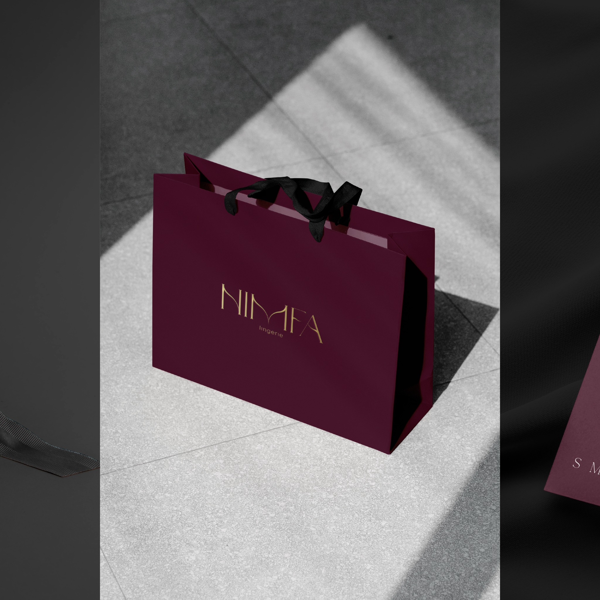 Elegancka, bordowa torba prezentowa z czarnymi uchwytami i złotym napisem 'NIMFA lingerie' leży na jasnoszarym tle w plamach światła.