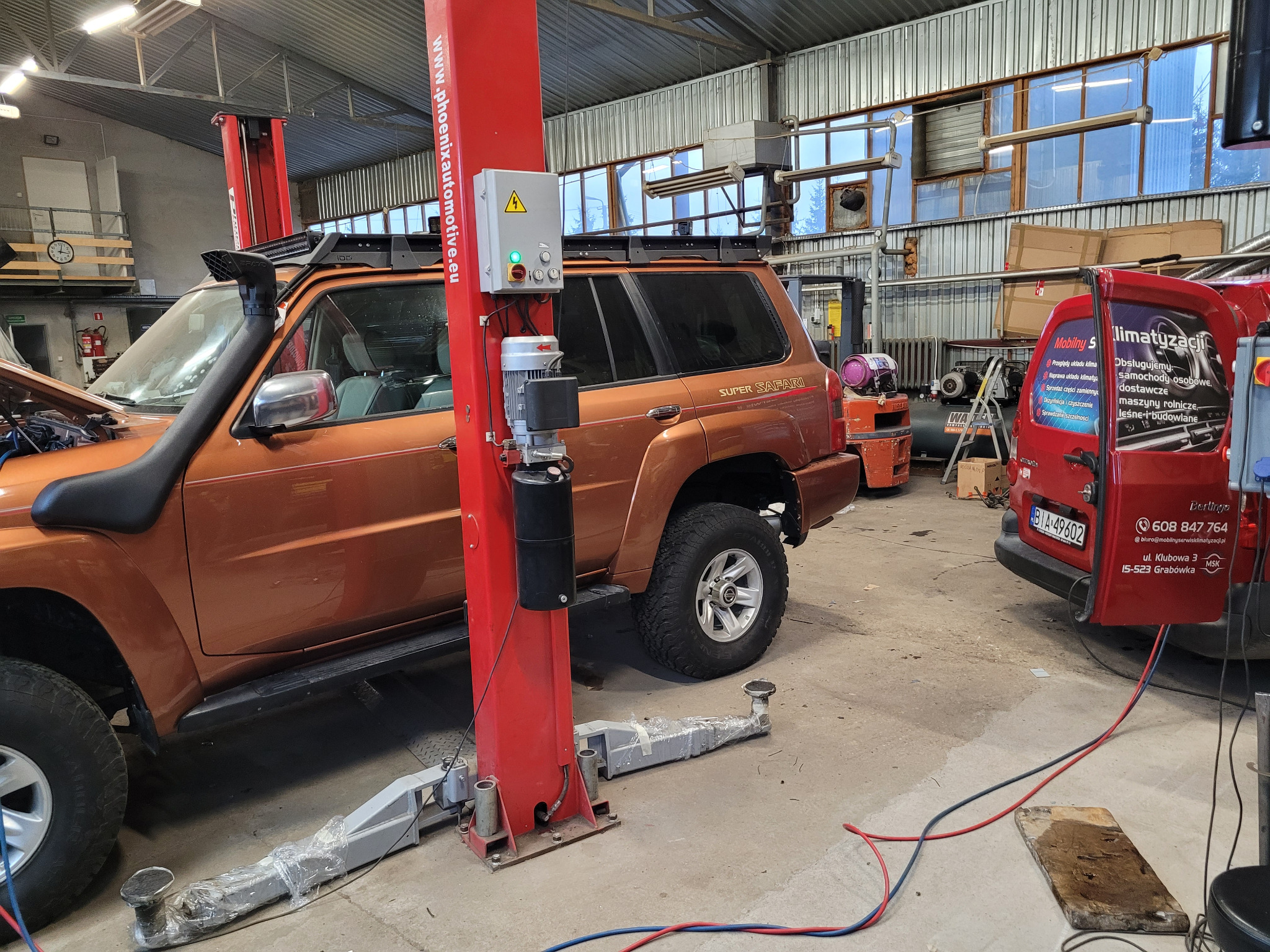 Pomarańczowy samochód terenowy na podnośniku w warsztacie, widoczny otwarty przód i podłączone przewody, obok czerwony samochód serwisowy z reklamą mobilnej klimatyzacji.