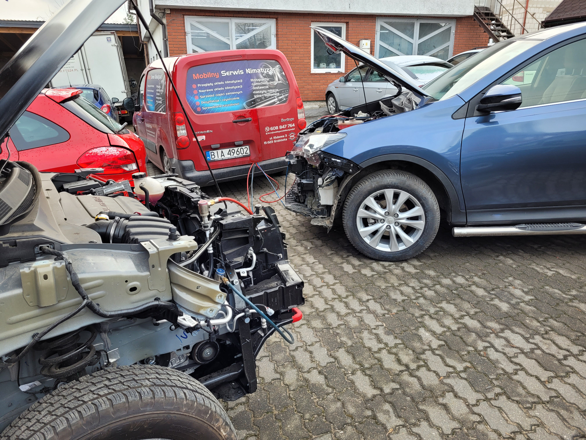 Warsztat samochodowy na zewnątrz: uszkodzony niebieski SUV z otwartą maską, czerwony samochód i czerwony van z reklamą serwisu klimatyzacji w tle, widoczne podłączone przewody i narzędzia.