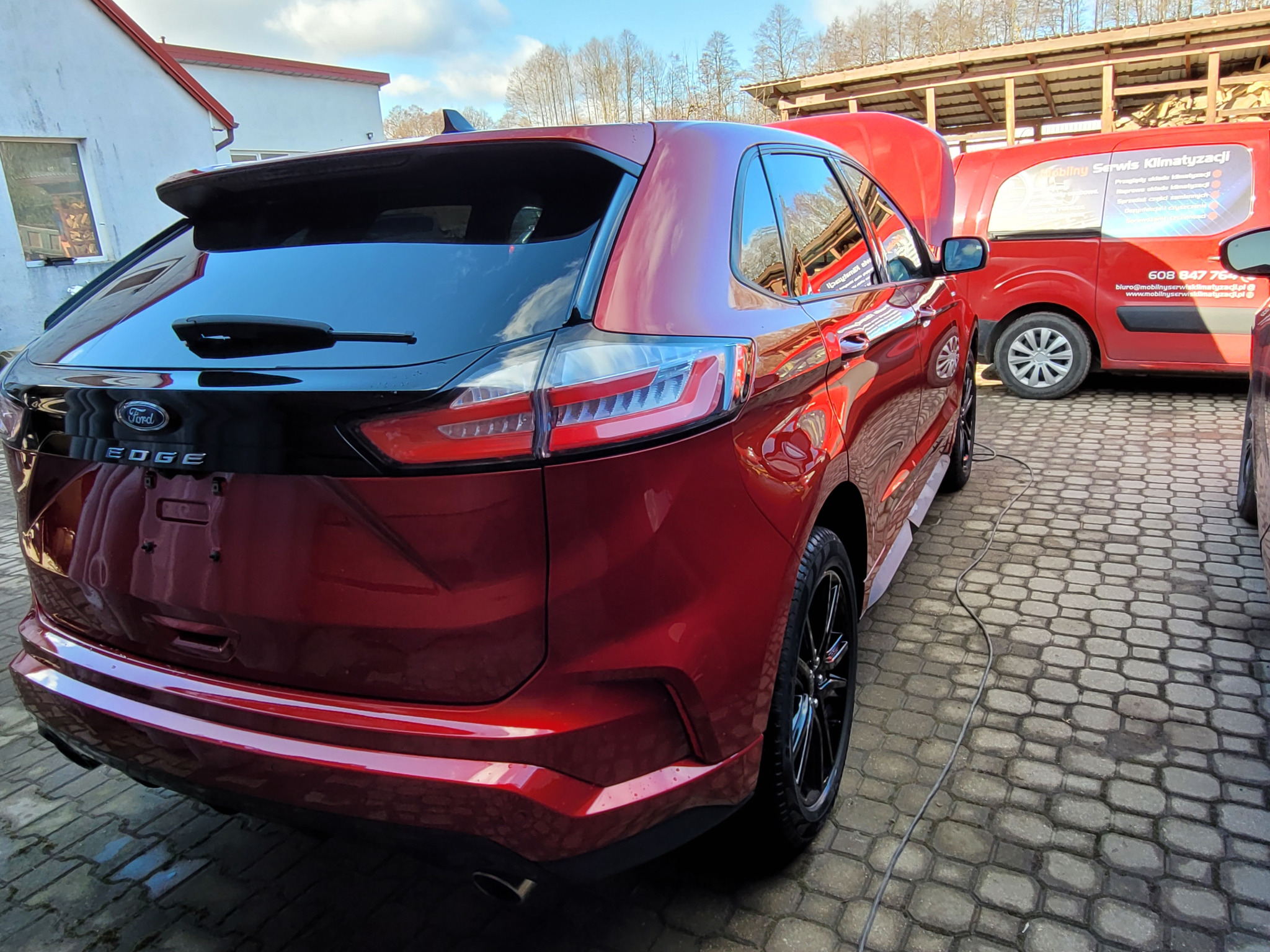 Czerwony Ford Edge z otwartą maską, zaparkowany obok czerwonego samochodu serwisowego z napisem 'Mobilny Serwis Klimatyzacji' na kostce brukowej.