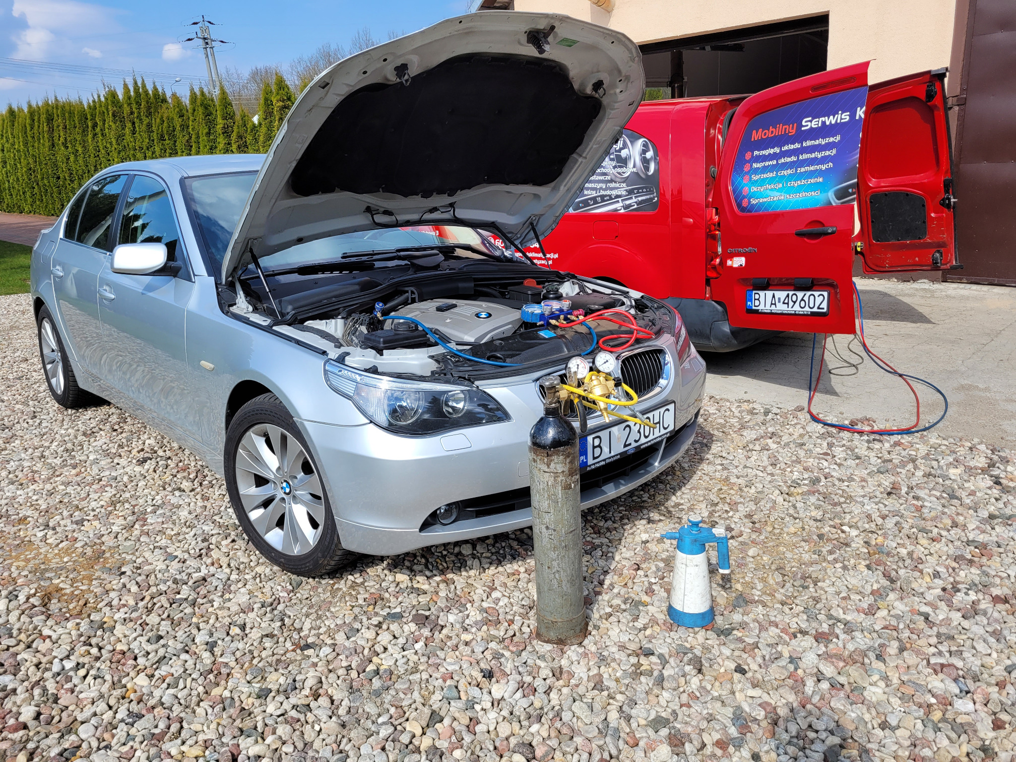 Srebrny samochód z otwartą maską podczas serwisu układu chłodzenia, widoczny podłączony sprzęt diagnostyczny i butla z czynnikiem chłodniczym, na tle czerwony samochód serwisowy z reklamą.