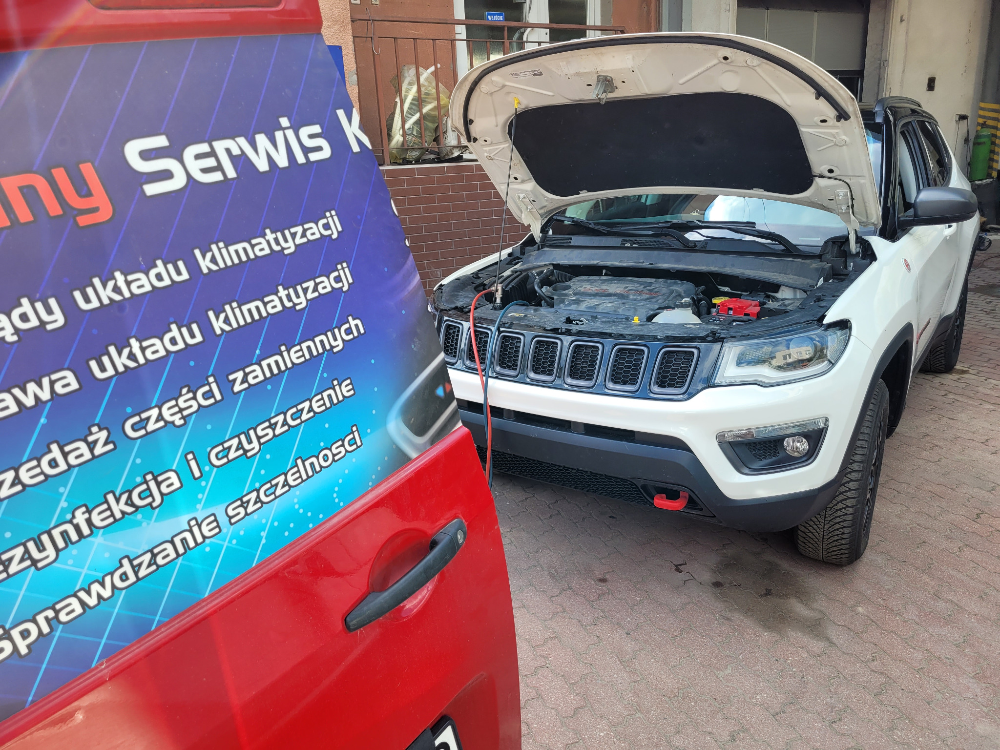 Biały samochód z otwartą maską, podłączony do maszyny serwisowej; widoczny plakat z ofertą usług, w tym odgrzybianie i sprawdzanie szczelności.