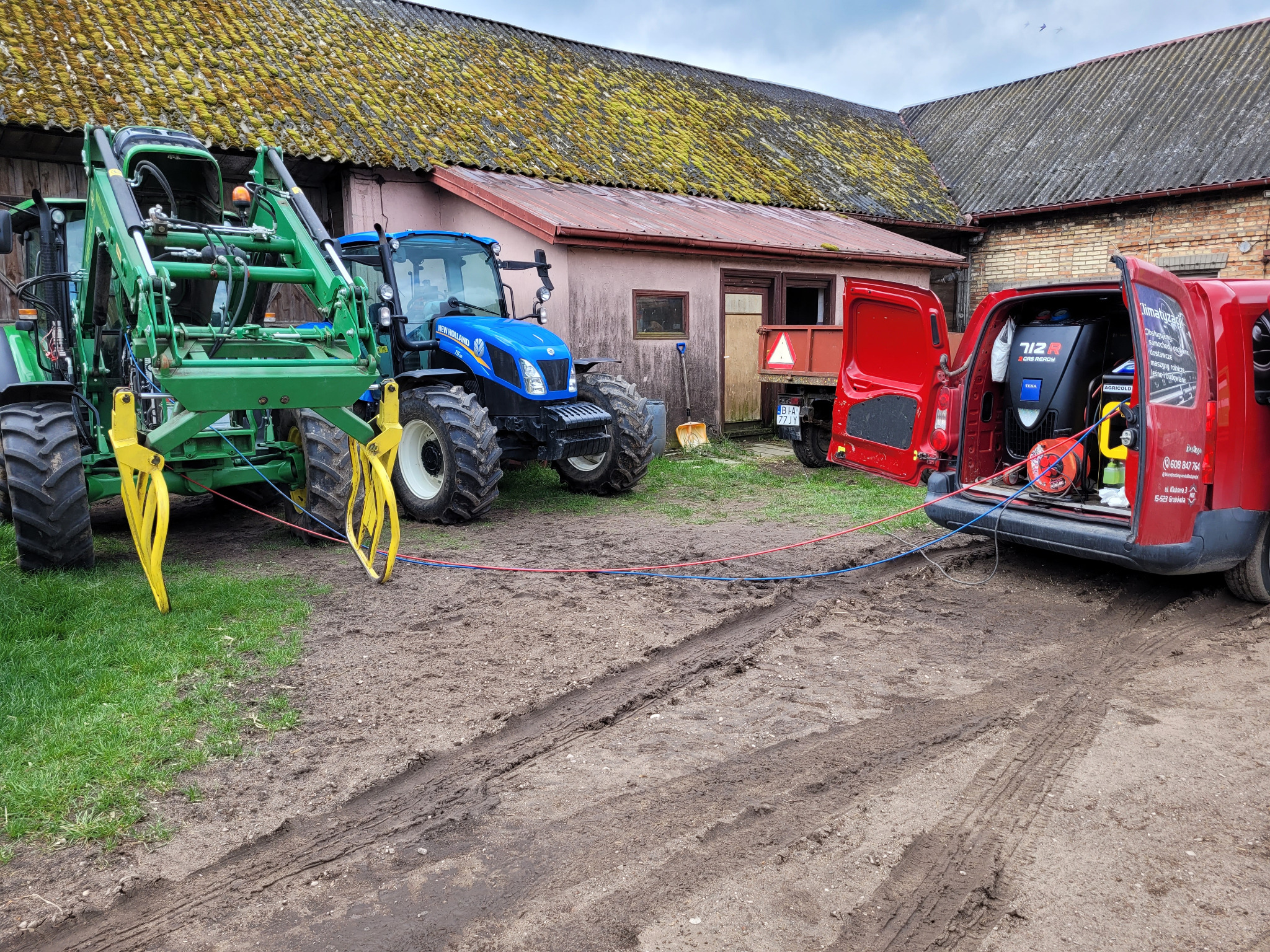 Serwis mobilny przy zielonym i niebieskim traktorze na błotnistym podwórku; czerwony van z otwartymi drzwiami bagażnika, z którego wychodzą przewody podłączone do traktorów; w tle budynek...