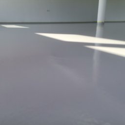 Posadzki żywiczne Podhale - Gładka, szara podłoga w hali przemysłowej z widocznymi promieniami słonecznymi wpadającymi przez okno i odbijającymi się od powierzchni. Kolumna pomalowana na biało stoi w rogu pomieszczenia.