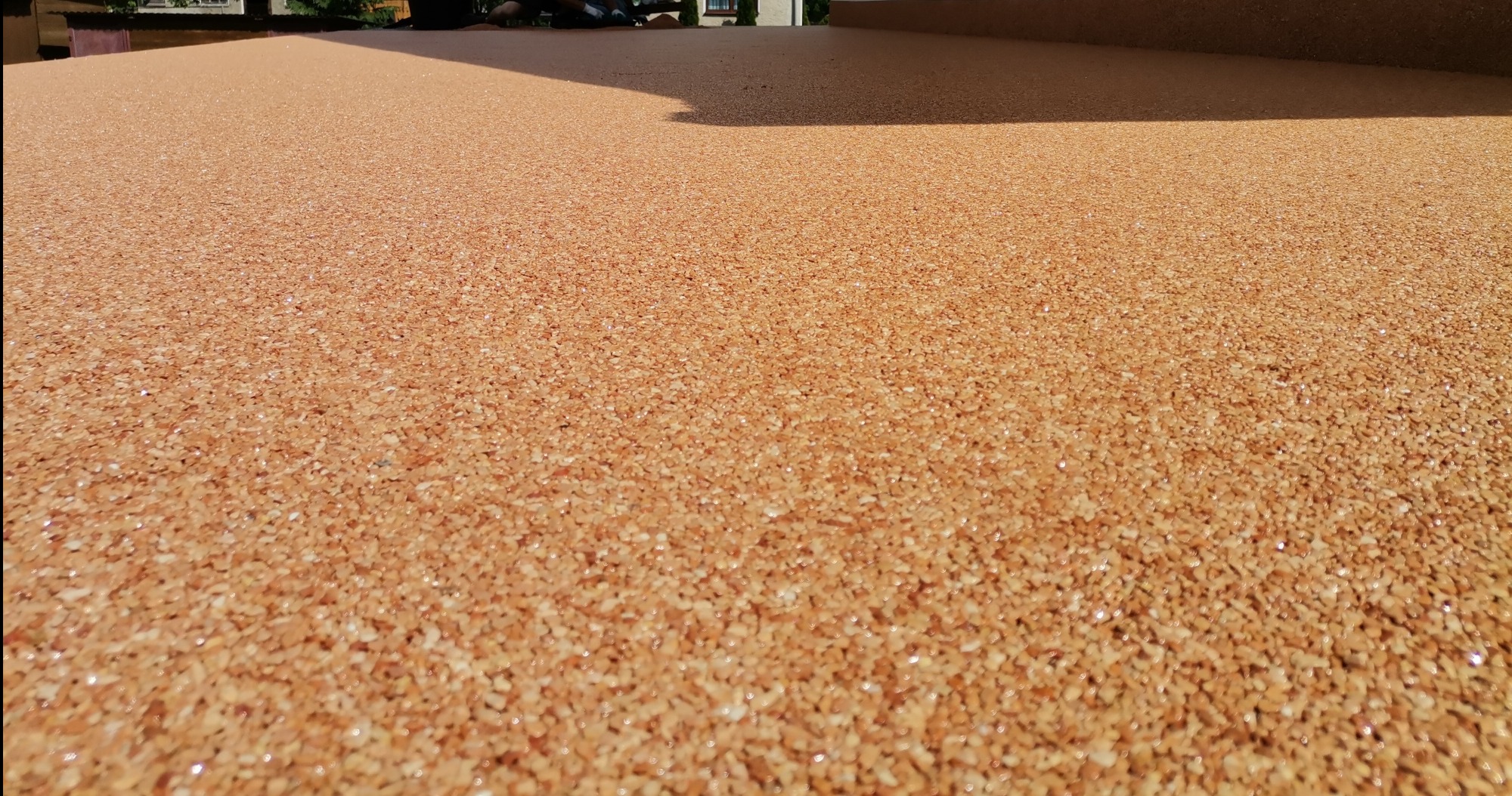 Jasnobrązowa posadzka żywiczna z widoczną strukturą drobnych kamyczków, lekko błyszcząca w promieniach słońca, z cieniem na górze