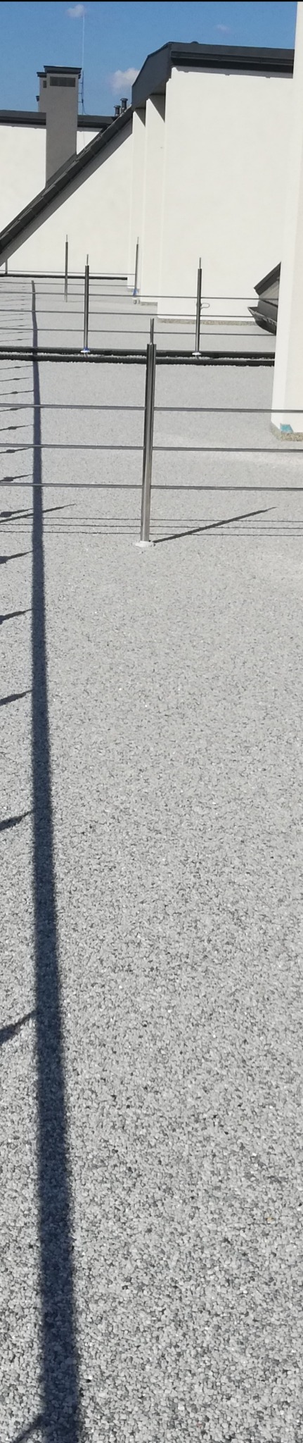 Szara, drobnoziarnista powierzchnia na dachu budynku z widocznymi metalowymi słupkami balustrady i fragmentami białych ścian w tle.