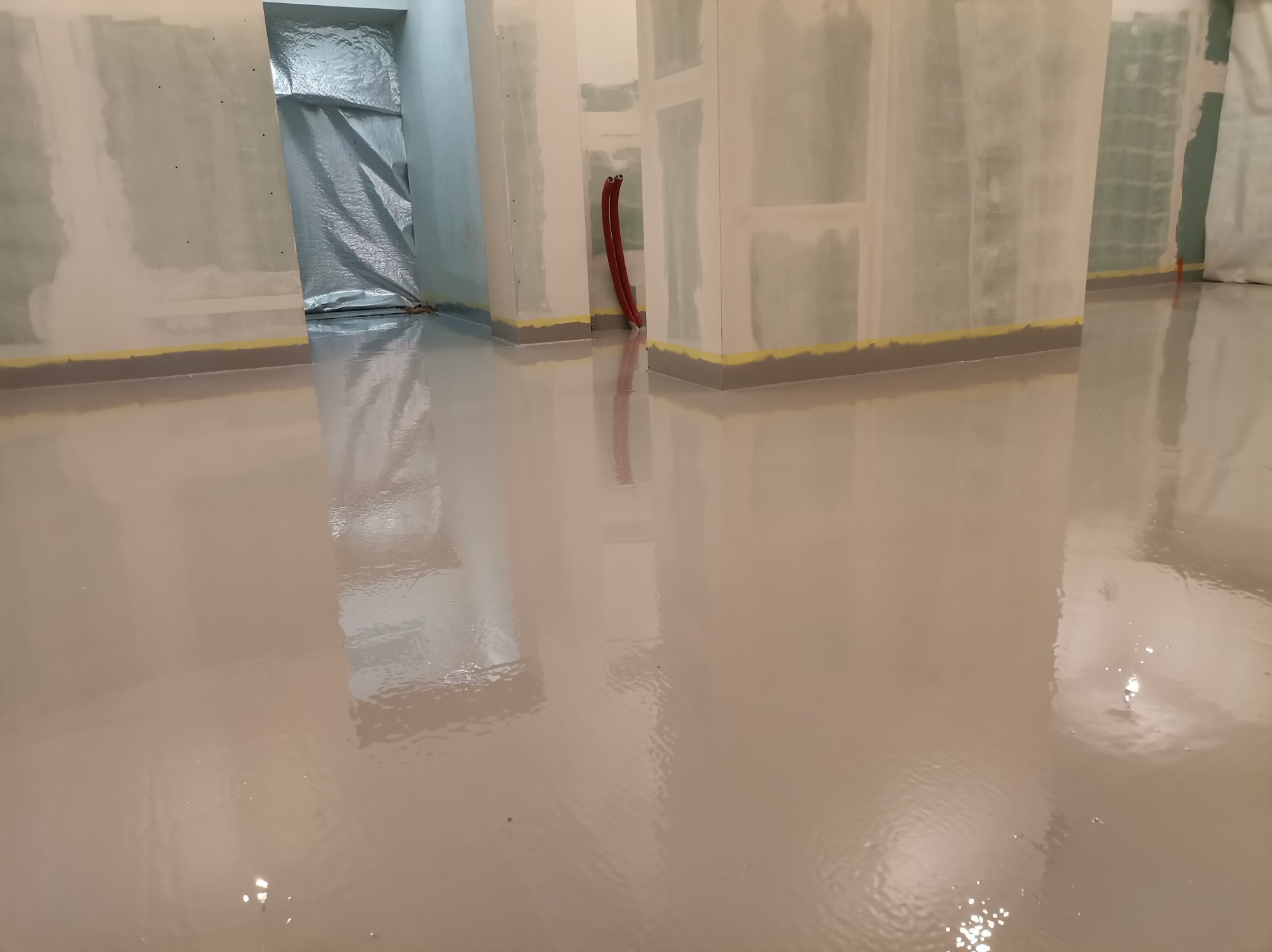 Świeżo wylana, błyszcząca, beżowa podłoga w surowym wnętrzu, zabezpieczona taśmą malarską przy ścianach, z widocznymi instalacjami i folią ochronną w przejściu.
