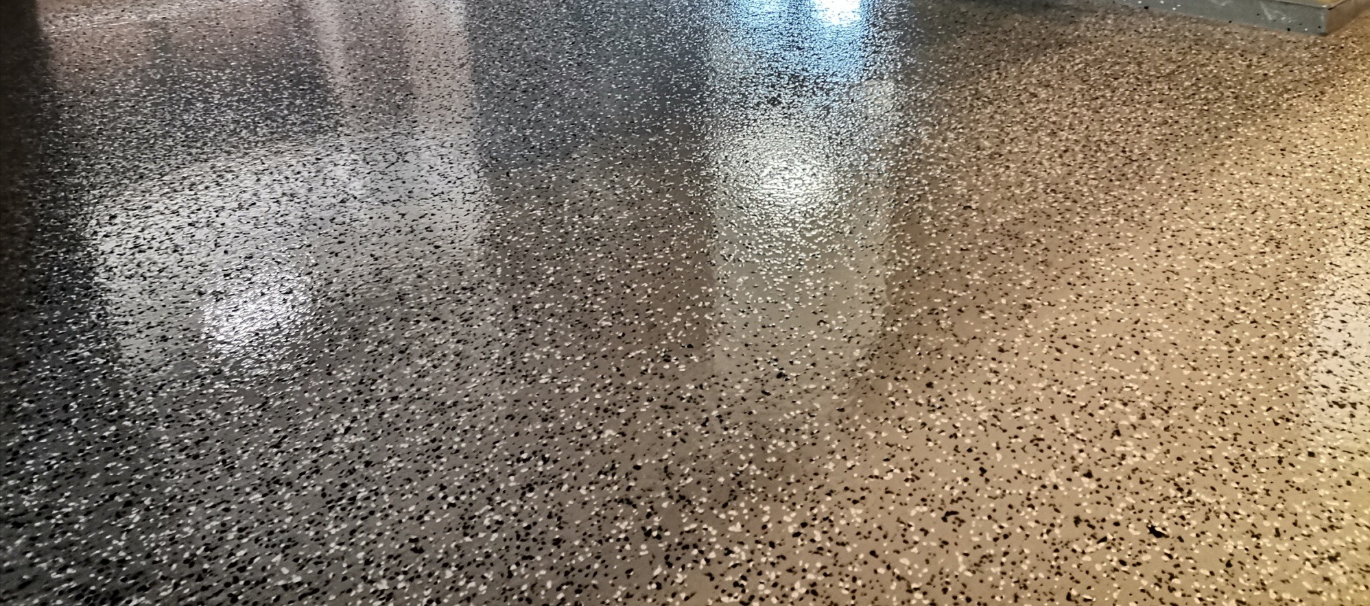 Błyszcząca, gładka powierzchnia podłogi z żywicy epoksydowej w odcieniach szarości z widocznymi drobnymi, wielobarwnymi elementami dekoracyjnymi.