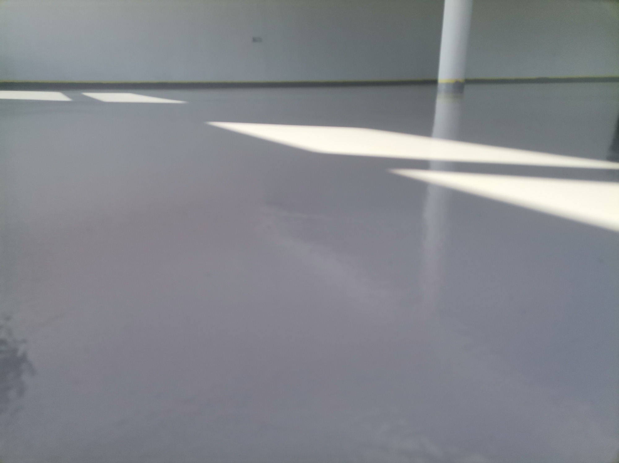 Gładka, szara podłoga w hali przemysłowej z widocznymi promieniami słonecznymi wpadającymi przez okno i odbijającymi się od powierzchni. Kolumna pomalowana na biało stoi w rogu pomieszczenia.