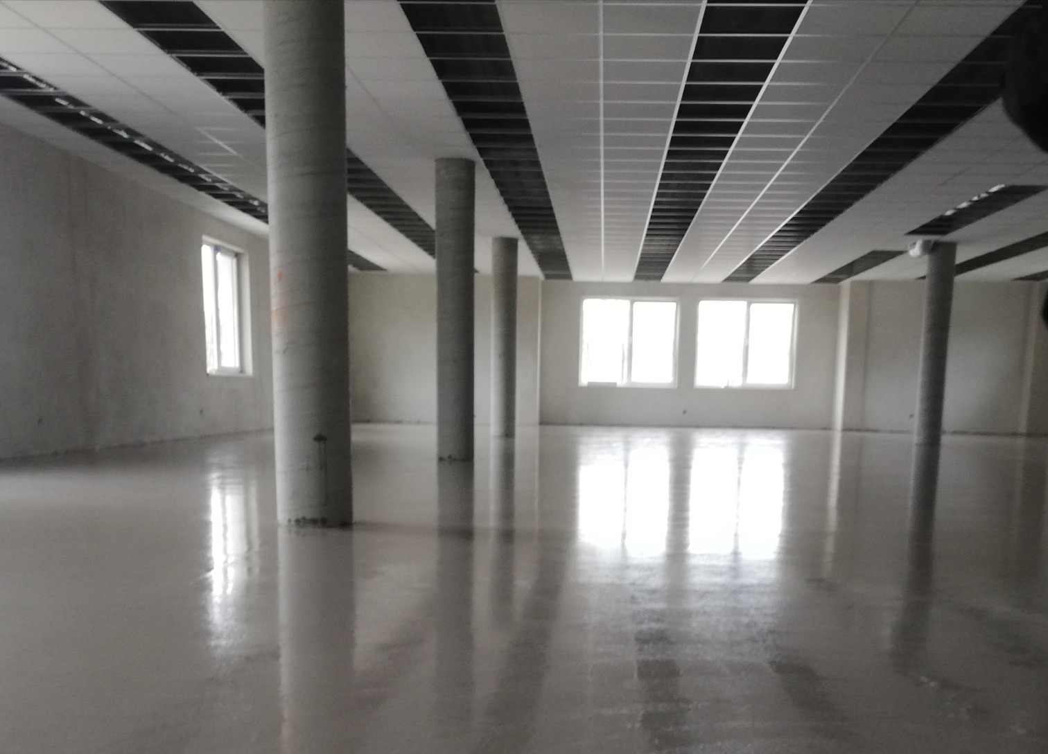 Wnętrze biurowe z wylaną, błyszczącą posadzką żywiczną, widoczne kolumny nośne i okna.