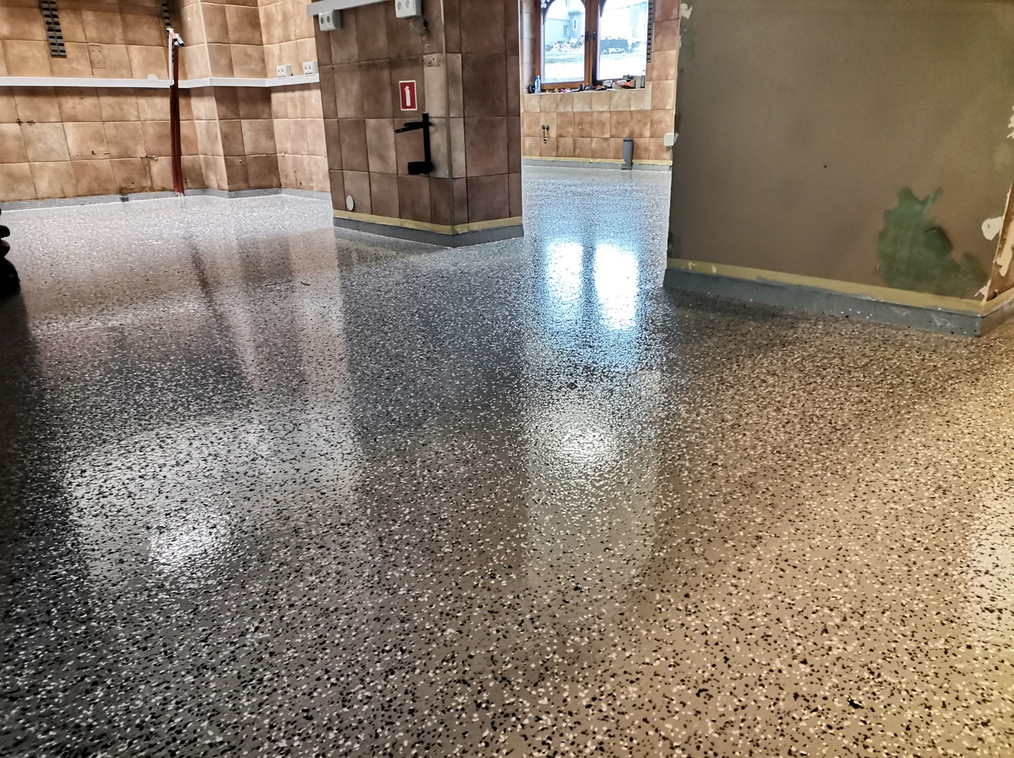 Błyszcząca, bezszwowa podłoga w kolorze szaro-beżowym z drobnymi, ciemnymi i jasnymi plamkami, odbijająca światło z okna i lamp, w tle fragment ściany z brązowych płytek.