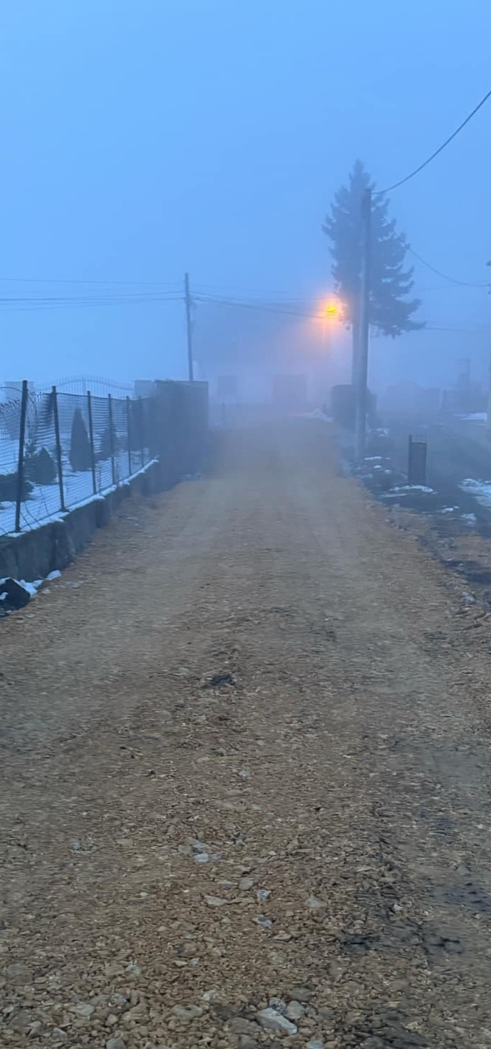 Droga gruntowa po robotach ziemnych, widoczna w oddali zabudowa w gęstej mgle, częściowo ośnieżona, oświetlona lampą uliczną.