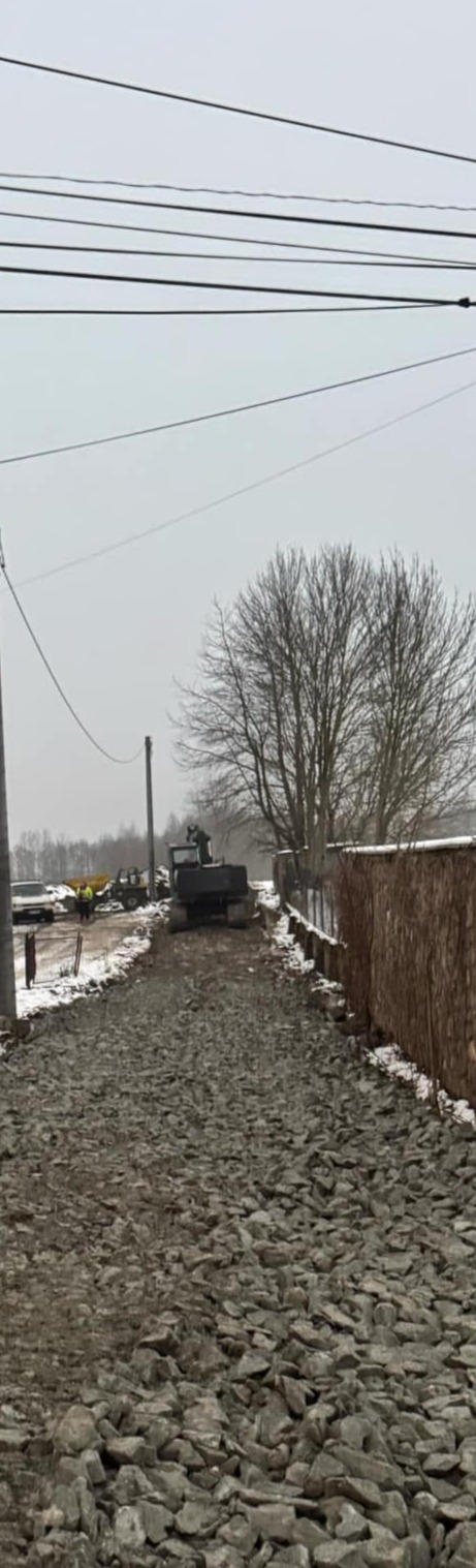 Ciężki sprzęt budowlany ugniata kamienistą drogę w pochmurny dzień, z widocznymi przewodami elektrycznymi nad głową i fragmentami śniegu na poboczu.